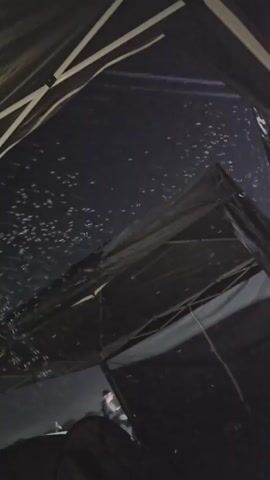 The video shows a dark, possibly nighttime scene, viewed from underneath a tent or canopy. The fabric of the canopy is black and has white support struts visible. Scattered across the upper surface of the canopy are numerous small, bright white specks. These specks appear to be illuminated by an external light source, possibly stars or distant lights, and are concentrated in the upper part of the frame. Below the canopy, other dark tent-like structures are visible, as well as a dimly lit background that suggests an outdoor environment. There is a slight blur to the image, indicating motion or low light conditions.