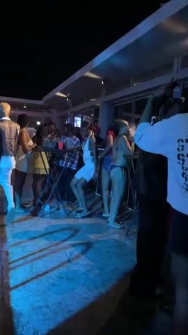 The video shows a group of people dancing at an outdoor party at night. Most of them are gathered around small tables with red plastic cups. One woman in a white dress is dancing in the center. Another woman is wearing a bikini and smoking something from a tall glass device. The area is lit with a blue light and has a low, flat roof with recessed lights.