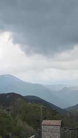 The video shows a mountain range landscape under a cloudy sky. The mountains are layered in the distance, fading into a light blue hue. In the foreground, there's a small stone building with a red-tiled roof, situated on a green hillside. A utility pole and a modern street lamp are visible near the building. The overall weather appears overcast, with diffused light filtering through the clouds. The scene is still and quiet.