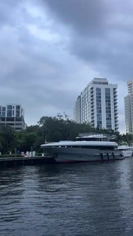 A video shows a grey yacht docked on the left side of a waterway. Behind the yacht, several tall white buildings are visible, with trees and palm trees in front of them. Another white boat, resembling a paddlewheel boat, is docked on the right side of the waterway. The sky is overcast with grey clouds. The water is dark and slightly choppy.