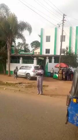 The video shows a street scene with vehicles passing by. Initially, a person is seen walking past a building, possibly a mosque. A white van with yellow stripes and a white sedan subsequently drive past. The street is lined with small shops selling clothes and other items. People are seen standing near the shops. The environment appears to be a roadside area in a developing region, with trees and foliage in the background.