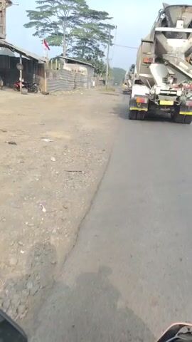 A video taken from a moving vehicle shows a concrete mixer truck driving down a two-lane paved road. The truck is in the right lane, moving away from the camera. On the left side of the road, there are several small, weathered buildings with corrugated metal siding, and a fence made of similar material. A motorcycle is parked near one of the buildings. A tall, leafy tree stands behind the buildings, and a utility pole is visible. In the distance, other vehicles, including a yellow truck and a blue truck, are also visible on the road. The sky is clear and the environment appears to be a rural or semi-industrial area.