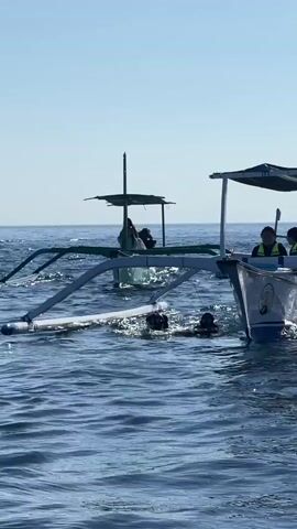 The video shows two outrigger boats in the ocean.  Some people in dark wetsuits are in the water, near the outrigger floats of the boats.  A man wearing sunglasses and a baseball cap observes the scene from within the front boat.  The sky is blue.