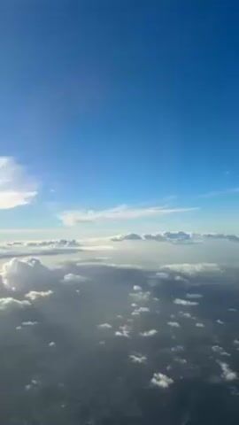 The video shows a high-altitude view from an airplane, with a clear blue sky at the top and a layer of fluffy white clouds below. The sun's rays pierce through the clouds, creating shadows and highlighting the landscape underneath. The perspective is from an airplane, looking downward, revealing some ground features beneath the clouds.