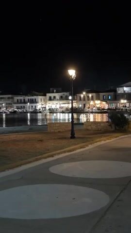 The video shows a night scene of a waterfront with buildings lining the shore. A streetlamp illuminates the foreground. The water reflects the lights from the buildings. There are people visible in the buildings and along the waterfront. The sky is dark.