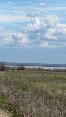 The video shows a car driving down a dirt road. The road is surrounded by a field of tall grass and shrubs. In the distance, there is a body of water and a cloudy sky. The video is shot from the perspective of the driver.