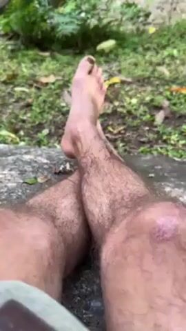 The video shows a man relaxing outdoors near a stream. The camera focuses on his bare feet and legs, which are hairy, and one knee appears to have a minor scrape or scab. He's sitting on a concrete surface covered in moss.  The background reveals a small, shallow stream flowing over rocks. There's green vegetation visible around the stream, including ferns and other plants.  The overall scene depicts a peaceful, natural environment.