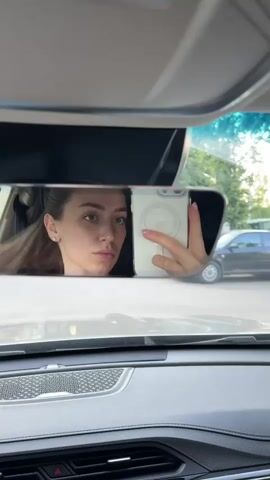 A young woman takes a selfie in a car's rearview mirror. She is looking into the mirror and holding her phone to take the picture. She makes a few different faces, including a wink and a kissy face. The car appears to be parked outdoors, as evidenced by the reflection of trees and a black car in the mirror.