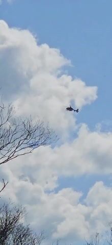 The video shows a helicopter flying against a blue sky with scattered white clouds. A bare tree branch is visible in the lower left of the frame. The helicopter is dark in color and appears to be moving away from the viewer.
