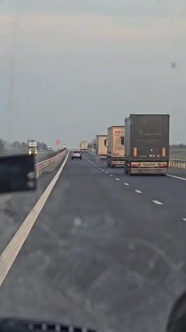 The video shows a view from inside a car driving on a multi-lane highway. The sky is overcast. In the distance, several large trucks are driving in the same direction. A smaller car is also visible on the same lane, further ahead. The road has white lane markings. A guardrail is visible on the left side of the road. Some signs can be seen in the distance. The car's dashboard is partially visible at the bottom of the frame.