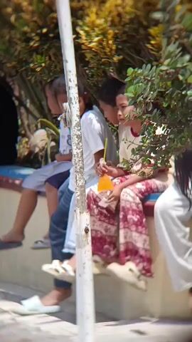 A group of four children are sitting on a low wall outdoors. The child on the far left is wearing a grey t-shirt and blue jeans, with dark hair. The child next to them is wearing a white t-shirt and dark pants, also with dark hair. The third child from the left is wearing a white t-shirt with a yellow straw in a cup of orange liquid, and patterned pants. The child on the far right is wearing a cream-colored shirt with red trim and patterned pants. Behind the children is a dense hedge of green and yellow foliage, and a white pole is visible in the foreground. The children are looking to their left.