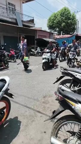 A vertical video depicts a street scene with multiple motorcycles parked and in motion. The timecode indicates the beginning of the video at 00:00 and the end at 00:04. In the foreground, several motorcycles are parked, with the rear wheel and fender of one prominently featured. The street is paved and appears dusty. In the middle ground, a white motorcycle is ridden by a person wearing a patterned shirt and shorts, moving from left to right. Further back, other motorcycles and people are visible. To the left, a red motorcycle is parked, and a person carrying a blue bag walks away from the camera. A circular red and white sign is visible on a pole. In the background, buildings line the street, and a large green tree stands out against a bright sky with scattered clouds. Other vehicles, including a truck, are also present in the distance. The overall impression is a busy street, possibly a market or commercial area, with a lot of motorcycle activity.