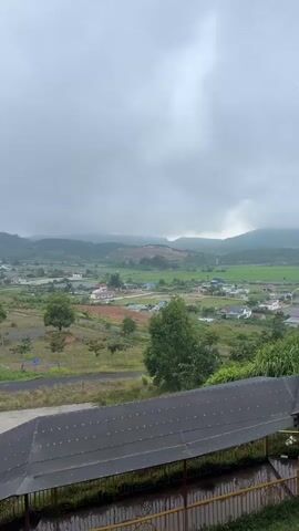 A vertical video shows a panoramic view of a valley landscape under a cloudy sky. In the foreground, a dark, corrugated roof structure extends across the bottom of the frame, partially obscuring the view. Below the roof, a metal railing and what appears to be water are visible. The middle ground features a grassy, somewhat uneven terrain with scattered trees, some with sparse foliage. A road curves through this area, and further in the distance, there are buildings and agricultural fields, likely rice paddies given their green color. The background is dominated by rolling hills and mountains shrouded in mist and the overcast sky, suggesting a humid or rainy climate. There are no people or animals visible in the video. The overall impression is one of a rural, possibly mountainous, landscape viewed from an elevated vantage point on an overcast day.