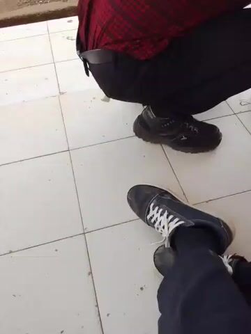 The video shows a person in a red and black plaid shirt crouching down and interacting with a young child. The child is wearing a gray t-shirt and blue shorts and is kneeling on the floor. In front of them is a toy truck, which appears to be orange and black. The person in the plaid shirt is holding the truck and seems to be showing it to the child. The child is looking at the truck and reaching out towards it. The floor is tiled with white and light gray tiles. In the background, there are plants, a building with pink pillars, and some furniture. The overall setting appears to be outdoors or in an open-air covered area, possibly a school or public space.