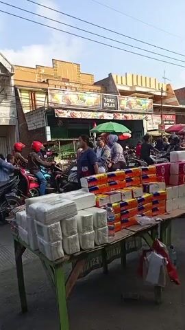 The video shows a busy street scene with several people riding on motorcycles and scooters. In the foreground, there is a stall selling paper products, with stacks of napkins and boxes of tissues displayed. Behind the stall, a group of people are gathered on motorcycles. Further in the background, there are buildings and shops, one of which has a sign that reads "TOKO IKHLAS". The sky is clear and blue. The overall impression is of a lively marketplace or street corner.