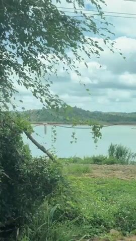 This is a landscape video of a large body of water with hills in the background. The water is a light blue-green color and there are some clouds in the sky. The foreground consists of dirt and some vegetation. The video appears to be filmed from a moving vehicle. There are power lines visible in the upper part of the frame. There are no people or animals visible in the video.