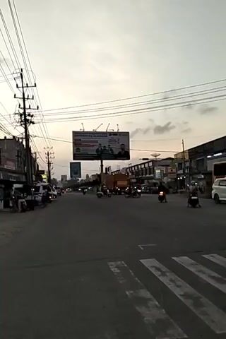 The video shows a street intersection in a city, likely in Southeast Asia. It is dusk, with the sky a mix of pale blue and orange. Power lines crisscross overhead. A large billboard for "Universitas Almuslim" dominates the view above the intersection. Several motorcycles and a few cars and vans are visible on the road. Some people are walking or standing on the sidewalks. The scene suggests a typical urban environment with moderate traffic at the end of the day.
