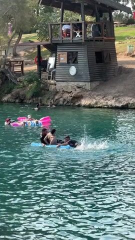 A group of people are enjoying a day at a lake. In the foreground, three young men are on a blue inflatable raft, splashing and playing in the water. One of the men is on his back, another is attempting to push himself up, and the third is on his stomach, facing forward. In the background, a few other individuals are swimming and floating on pink and white inflatable rings. Behind them, a wooden lifeguard tower with a thatched roof stands on the shore. The tower has multiple levels and windows, with people visible on the upper deck. The lake is a clear, turquoise color, with ripples on the surface. The surrounding environment is lush and green, with trees and a grassy slope leading up to the tower. The sun is shining, casting a warm glow on the scene.