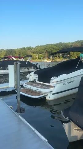 The video shows a view from a dock at a marina. Several boats are docked on both sides of the dock. The boats are of various sizes and types, including pontoon boats and a speed boat. Some of the boats are covered with tarps. The water in the marina is calm and reflects the sky and the boats. The sky is clear and blue. There are trees in the background on the shore. The sun appears to be low in the sky, suggesting it is either early morning or late afternoon.
