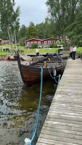 The video begins with a close-up of the front of a wooden Viking-style boat docked at a wooden pier. The boat has a dragon head carved into its bow. The water is choppy. A blue rope secures the boat to the pier. As the camera pans to the right, the pier and the boat continue to be visible, with people standing on the pier and in the background. Beyond the pier, there is a grassy area with several red buildings, some with white trim. Trees, including tall birch trees, are scattered throughout the scene. In the background, a group of people are gathered on the grass, and another person stands near one of the buildings. The sky is overcast. The camera then pans further right, revealing more of the water and the edge of the pier. The red buildings and trees remain in the background. The video ends with a shot of the water and the pier from a slightly lower angle.