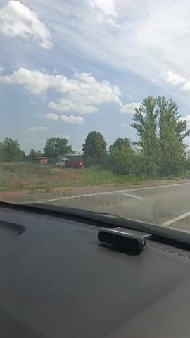 The video shows a view from inside a car, looking out the windshield. The car is driving on a paved road, possibly a highway. On the right side of the road, there is a grassy median and then more road. On the left side of the road, there are trees, bushes, and some buildings or structures in the distance. The sky is blue with white clouds. A small black device, likely a dashcam, is visible on the dashboard. The video captures the car moving forward, with scenery passing by. There are a few other vehicles visible in the distance on the road.