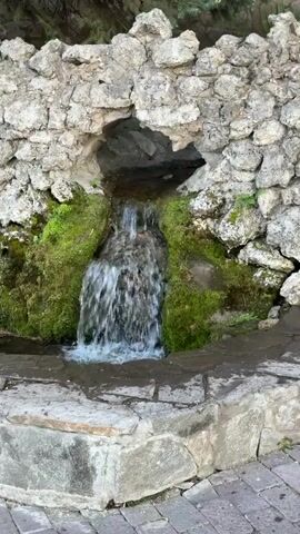 The video shows a small artificial waterfall and fountain feature. The water flows from a hole in a rock structure, over moss, and into a stone basin. The fountain is constructed from rough-hewn stones and bricks and appears to be set in a public space, as evidenced by the brick pavers visible at the bottom of the frame.  A pine tree is visible in the background.