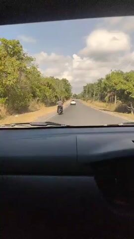 The video shows a view from inside a car looking out the windshield at a two-lane road. The road is lined with trees and vegetation on both sides. A person is riding a scooter in the right lane of the road, heading away from the camera. Behind the scooter, there are two cars in the same lane. The sky is blue with some white clouds. The dashboard of the car is visible in the foreground. The overall impression is of a sunny day on a rural road.