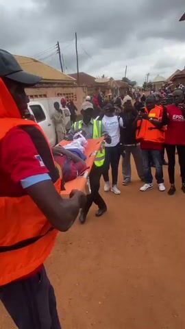 The video captures a scene in what appears to be a village setting. A person in an orange life vest is walking away from the camera, turning to look back. In the background, a crowd of people has gathered. A stretcher, carrying an individual lying on their back, is being carried by several individuals wearing safety vests and orange life vests. The person on the stretcher is wearing a red and blue striped shirt and has a white bandage wrapped around their head. The environment consists of a dirt road lined with buildings, some appearing to be made of mud or brick, and a cloudy sky. Several people in the crowd are holding up phones, seemingly recording the event.