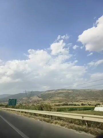 The video shows a view from a moving vehicle on a highway. The road curves to the left. To the right of the road is a metal guardrail, beyond which is a field with tall dry grass and some green vegetation. In the distance, there are rolling hills covered in greenery and trees. A large electricity pylon with power lines stretches across the landscape. The sky is blue with scattered white clouds. A green road sign with white arrows indicating lanes is visible on the right side of the road. The overall scene depicts a rural landscape under a clear sky.