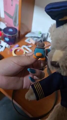A hand with long turquoise nails holds a small clay figure of Perry the Platypus, made of teal, brown, and orange clay. The hand then turns the clay figure to face a teddy bear dressed in a navy blue uniform with gold stripes. The teddy bear has a navy blue hat. The background is a wooden desk cluttered with various objects, including a colorful tin, scissors, a hair straightener box, and various other items. The camera movement is shaky and follows the hand's movement.