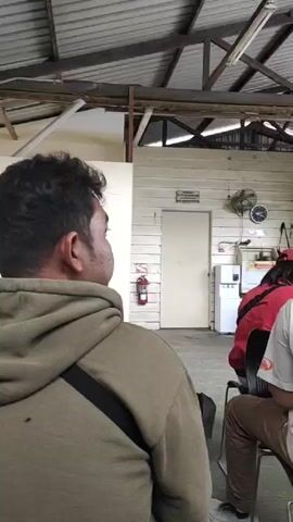 The video shows an indoor scene with several people sitting on chairs, facing away from the camera. The environment appears to be a waiting area, with wooden walls and a metal roof structure. In the background, there is a white door, a water dispenser, a fan, and a fire extinguisher. A poster is displayed on the wall. The people are casually dressed, and some are wearing hats. The lighting suggests it is daytime, possibly with natural light entering from an unseen source. The atmosphere seems calm, with no obvious activity or interaction captured.