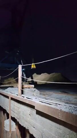 The video shows a pile of what appears to be sand, possibly gravel, being loaded by a crane at night. The crane's bucket picks up material from the mound and drops it to the side. A concrete structure is visible in the foreground, and a blue crane arm can be seen in the background. The lighting is dim, and the sky is dark, suggesting the video was recorded in a nighttime environment.