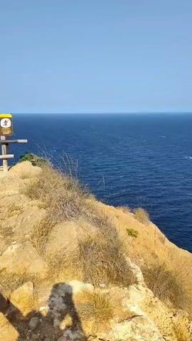 The video shows a view of the ocean from a rocky cliff. The sky is clear and blue, and the water is a deep blue with small waves. There is dry grass and a few green bushes growing on the cliff. The sun is shining, casting shadows on the rocks. The camera is moving slightly, panning across the scene. There is a shadow of a person visible in the bottom left corner of the frame.