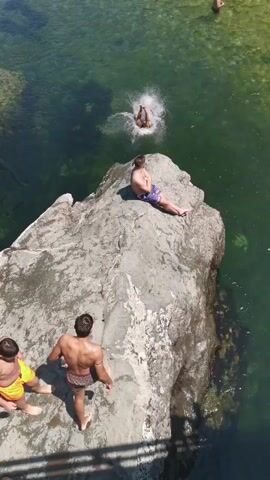 The video shows a group of people at a rocky body of water on a sunny day. Three young men are visible on a large rock in the foreground. One man, wearing purple swim trunks, is sitting on the edge of the rock. Two other men are standing on the rock, facing away from the camera. One of them is wearing yellow swim trunks and the other is wearing patterned swim trunks. In the background, a man is jumping into the water from a higher point on the rock, creating a splash. Another person is in the water swimming. The water is clear and green, and the surrounding area appears to be natural and undeveloped.