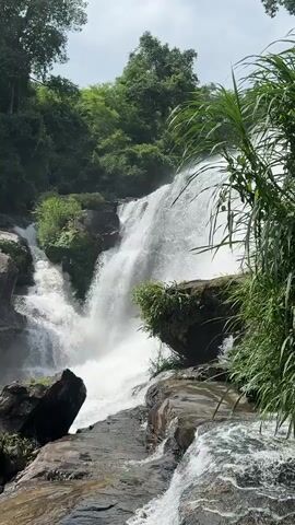 The video shows a natural scene with a waterfall. The waterfall is the main focus, with water cascading down a rocky cliff face. Lush green vegetation surrounds the waterfall, including trees and plants. The water is white and frothy as it falls, creating a misty spray. Rocks of various sizes are scattered around the base of the waterfall and in the foreground, some of which are wet from the water. The sky is visible in the background, with some clouds. The overall impression is one of a serene and natural environment.