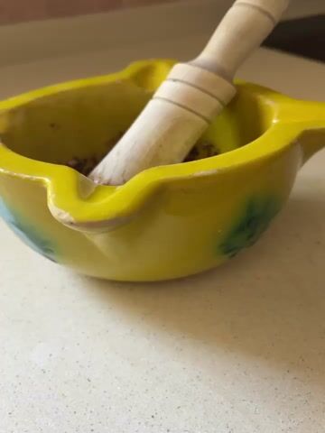 A close-up video shows a yellow ceramic mortar and wooden pestle. The mortar contains a dark brown paste-like mixture with visible herbs and possibly garlic. The pestle, covered in the same brown mixture, sits partially submerged in the paste. The mortar is resting on a light-colored countertop.