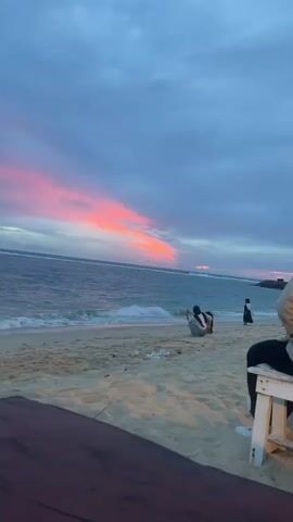 A video clip opens on a beach at dusk. The sky is a muted blue and gray, with streaks of vibrant pink and orange from the setting sun. Gentle waves lap at the sandy shore. In the mid-ground, a person is crouched on the sand, appearing to take a photo of the water. Another person stands further down the beach, facing the ocean. In the foreground, the edge of a wooden table and a white chair with someone sitting on it are visible. The overall impression is a peaceful, serene moment at the end of the day.