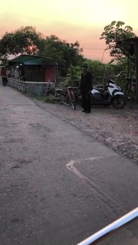 The video shows a first-person perspective of someone on a motorbike. The camera is positioned to capture the rider's reflection in the motorbike's side mirror and the road ahead. In the initial shot, an elderly man in a hat and dark clothing stands by a bicycle and a scooter on a dirt road. The environment appears to be a rural or semi-rural area with vegetation and structures. As the motorbike moves forward, the scene changes to show a paved road with a small market stall on the right side. A person in a red shirt is seen walking away from the stall. The sun is setting, casting a warm glow, and the sky is tinged with orange and pink. The video captures everyday life in what seems to be a developing country.