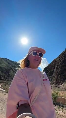 The video shows a woman in a pink outfit walking in a mountainous area with a sunny sky. She is wearing a pink cap and white sunglasses. The mountains in the background appear rocky and dry, with some sparse vegetation. The terrain she's walking on is a dirt path. There's a small canal-like structure next to the path. The woman seems to be recording herself as she walks and takes in the view.