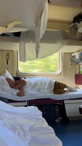 A person is sleeping in a bunk on a train. The person is covered by white sheets and is lying on their side, facing the window. The window shows trees and greenery passing by, indicating the train is in motion. The train compartment has multiple bunks, with shelves and storage above. There is a pillow under the person's head and a bag underneath the bunk.