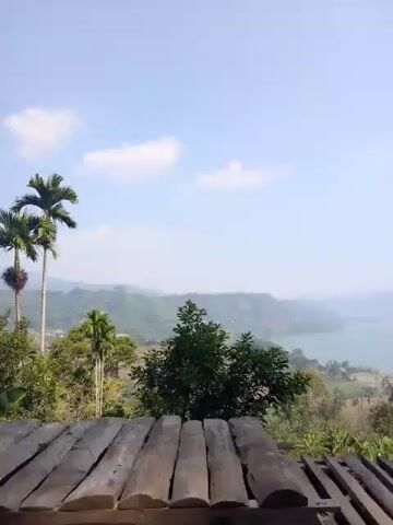 The video shows a view of a lake and rolling hills from a wooden platform. The sky is bright and clear with a few clouds. Trees and foliage are visible in the foreground and middle ground. The camera pans slightly to the right, revealing more of the landscape.