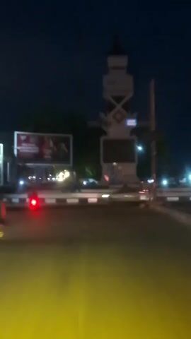 The video shows a nighttime scene in what appears to be a city. In the foreground, a motorcycle with its red taillight illuminated is moving from left to right across the frame. The road surface is illuminated with a yellow glow, possibly from streetlights or the motorcycle's headlight. In the middle ground, there is a roundabout with a monument or structure in the center. The monument is lit and has a dark rectangular panel on its front. Two large billboards are visible behind the roundabout. The billboard on the left has bright lighting and displays an image and text, including the words "A MAN TASTE". The billboard on the right is also lit and displays a red advertisement. Orange streetlights are visible on poles around the roundabout, and a traffic signal is also present. The background is dark, indicating it is nighttime, with some trees or foliage visible behind the billboards. The overall impression is a busy urban intersection at night.