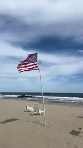The video shows an American flag, tattered and torn, waving on a pole on a sandy beach with the ocean in the background under a sky with scattered clouds.  Two beach chairs are positioned behind the flagpole, partially obscured by its shadow. The scene is static with the flag waving gently in the breeze.
