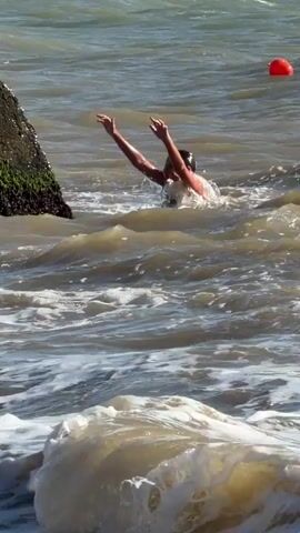 A person is struggling in rough, choppy water near a rocky structure. They appear to be reaching out towards the rocks, possibly for assistance or to gain a better grip. The water is a murky brown color with white foam from the waves. A red buoy is visible in the distance. The overall scene suggests a challenging situation for the individual in the water.