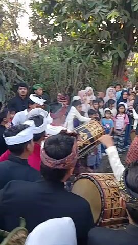 A group of people, some in traditional attire, are gathered outdoors in what appears to be a village setting. Several individuals are playing drums, which are elaborately decorated. A crowd of onlookers, including children and adults, watches the performance. The environment is characterized by trees and foliage, suggesting an open, possibly rural, space. The overall atmosphere seems festive, with participants and observers engaged in the event.
