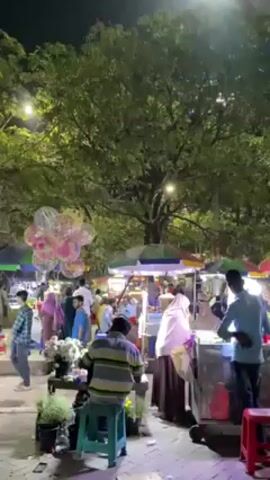 The video shows a bustling night market scene in what appears to be a city park. Many people are milling around, browsing various vendors with colorful umbrellas and carts. There are food stalls and a balloon vendor, and the area is illuminated by streetlights and lights from nearby buildings. In the background, a monument with red and white elements is visible, suggesting the location may have historical or cultural significance. The overall atmosphere is lively and crowded.