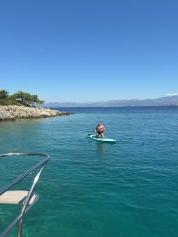 A person is seen stand-up paddleboarding (SUP) in clear, turquoise water under a bright, clear blue sky. The video is shot from a boat, with a chrome railing visible in the foreground.

The paddler, wearing a dark swimsuit, is initially kneeling on a light blue SUP board. They are holding a paddle and making strokes in the calm water. After a few seconds, they transition from a kneeling position to a standing position on the board, continuing to paddle forward.

The scene features a rocky coastline to the left with sparse green trees. In the distance, across the open water, a range of mountains is visible under the clear sky. The water is exceptionally clear, revealing the seabed near the boat. The overall impression is one of a serene and sunny day, perfect for watersports. No text is visible in the video.
