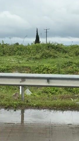 From a moving vehicle, a roadside view shows a rainy landscape with deteriorating industrial sites. Buildings range from incomplete concrete structures to weathered metal sheds. Debris such as stone piles and machinery clutter the area. The weather is overcast.