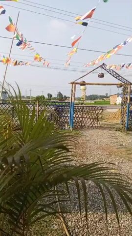 The video shows an outdoor scene with strings of international flags strung across the sky. In the foreground, a person is walking away from the camera down a gravel path. The person is wearing a dark shirt and pants. Behind the person, there is a bamboo fence with blue posts, and a small structure that appears to be a covered seating area. There are motorcycles parked behind the fence. In the distance, there are buildings and fields, and a water tower. The sky is clear and blue.