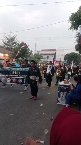 The video shows a procession on a street. Several people are marching in a line. Some are carrying flags, including Indonesian flags. The people are wearing black outfits, some with sashes. There is a banner that reads "PERGURUAN PECAT SILAT INDONESIA (IPSI)". In the background, there are buildings and trees. On the right side of the street, there are spectators seated, and a display box with the words "YOGURT STICK". The sky is overcast.
